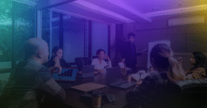 Team meeting in a conference room, with one person presenting to colleagues seated around a table with laptops and tablets.