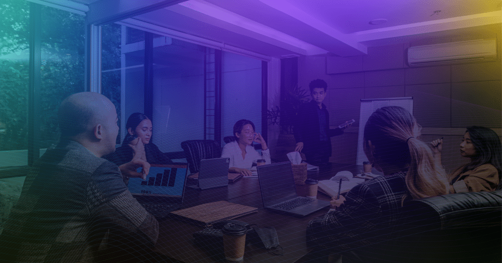 Team meeting in a conference room, with one person presenting to colleagues seated around a table with laptops and tablets.